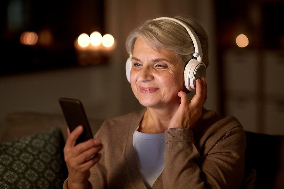 Äldre kvinna som sitter med hörlurar och tittar på en smartphone
